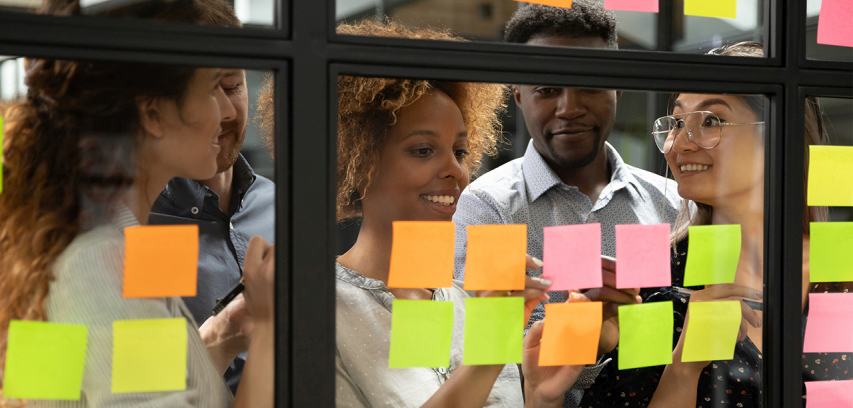 Business-agility-pillar-agile_orgs_photo_manifesto_scrum_meeting A group of colleagues smiles and collaborates with sticky notes on a glass wall